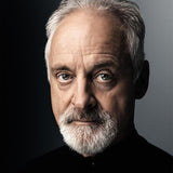 An older man with short white hair and a beard gazes directly at the camera, light eyes twinkling and a subtle smile gracing his face. He wears a black shirt against a gradient backdrop of black to gray, perfectly embodying the essence of Stravinsky: The Soldiers Tale by the London Symphony Orchestra.