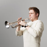 A person in a white sweater plays a silver trumpet against a gray background, focused on their instrument. Their wristwatch is visible. They embody the spirit of Stravinsky: The Soldiers Tale, from the London Symphony Orchestra.