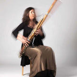 A woman in a long brown skirt and black top is seated on a chair playing a bassoon. Her hands and the instrument are captured in movement against a plain, light-colored background.