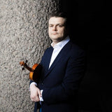 A person in a suit leans against a textured wall with Stravinsky: The Soldiers Tale by the London Symphony Orchestra, holding a violin. They look directly at the camera with a neutral expression against a dim background.